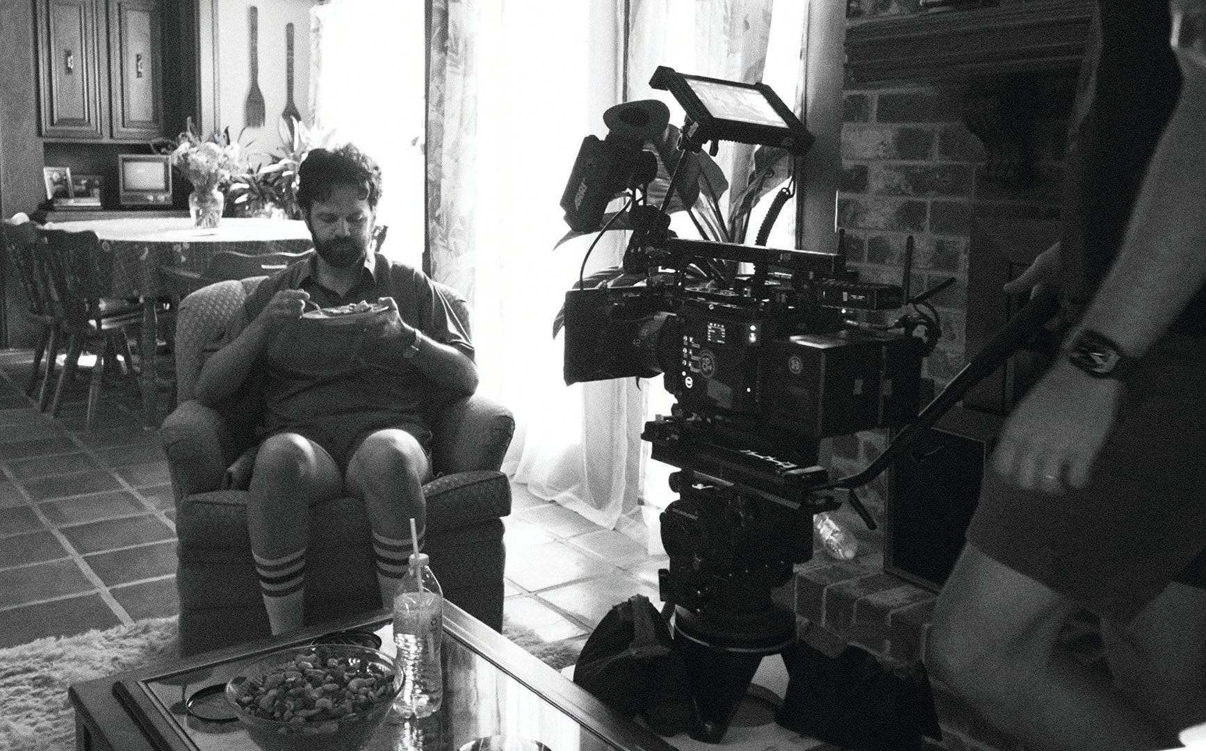 On set in black and white: a man in the living room armchair, the camera rig in the foreground