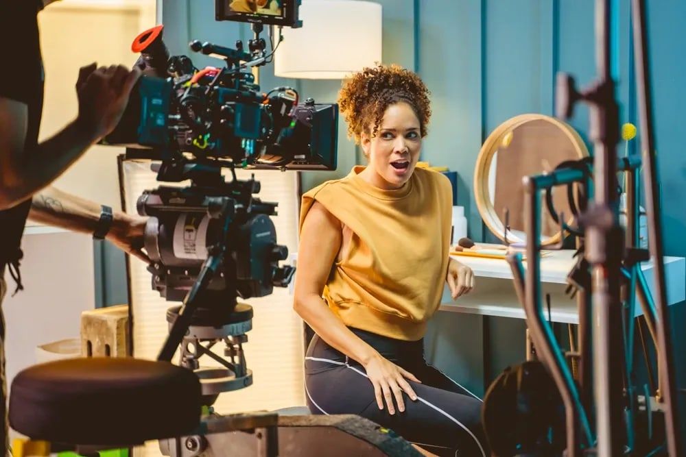 The camera crew rolls on a fitness spot as its star, in a mustard top, turns from her vanity mid-line