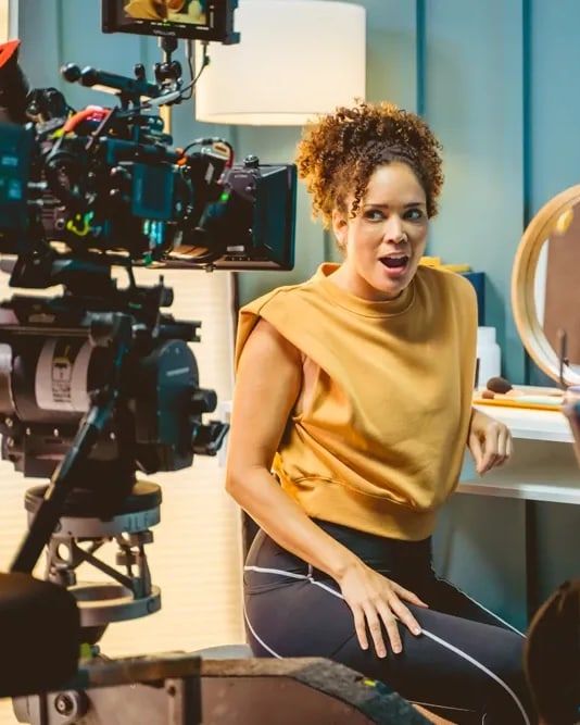The camera crew rolls on a fitness spot as its star, in a mustard top, turns from her vanity mid-line