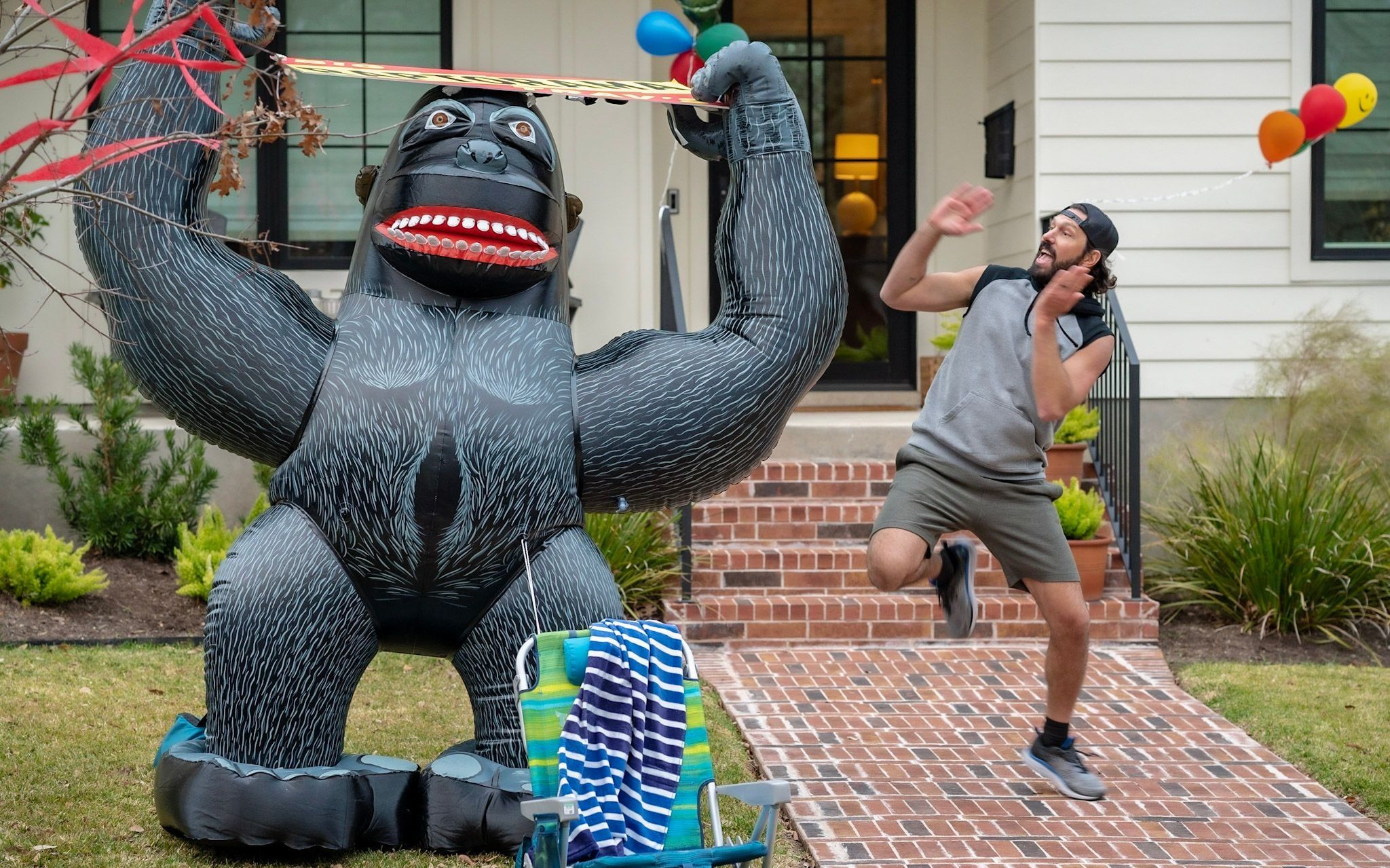 A giant inflatable gorilla holds a banner over a front yard while the talent leaps back in mock fright
