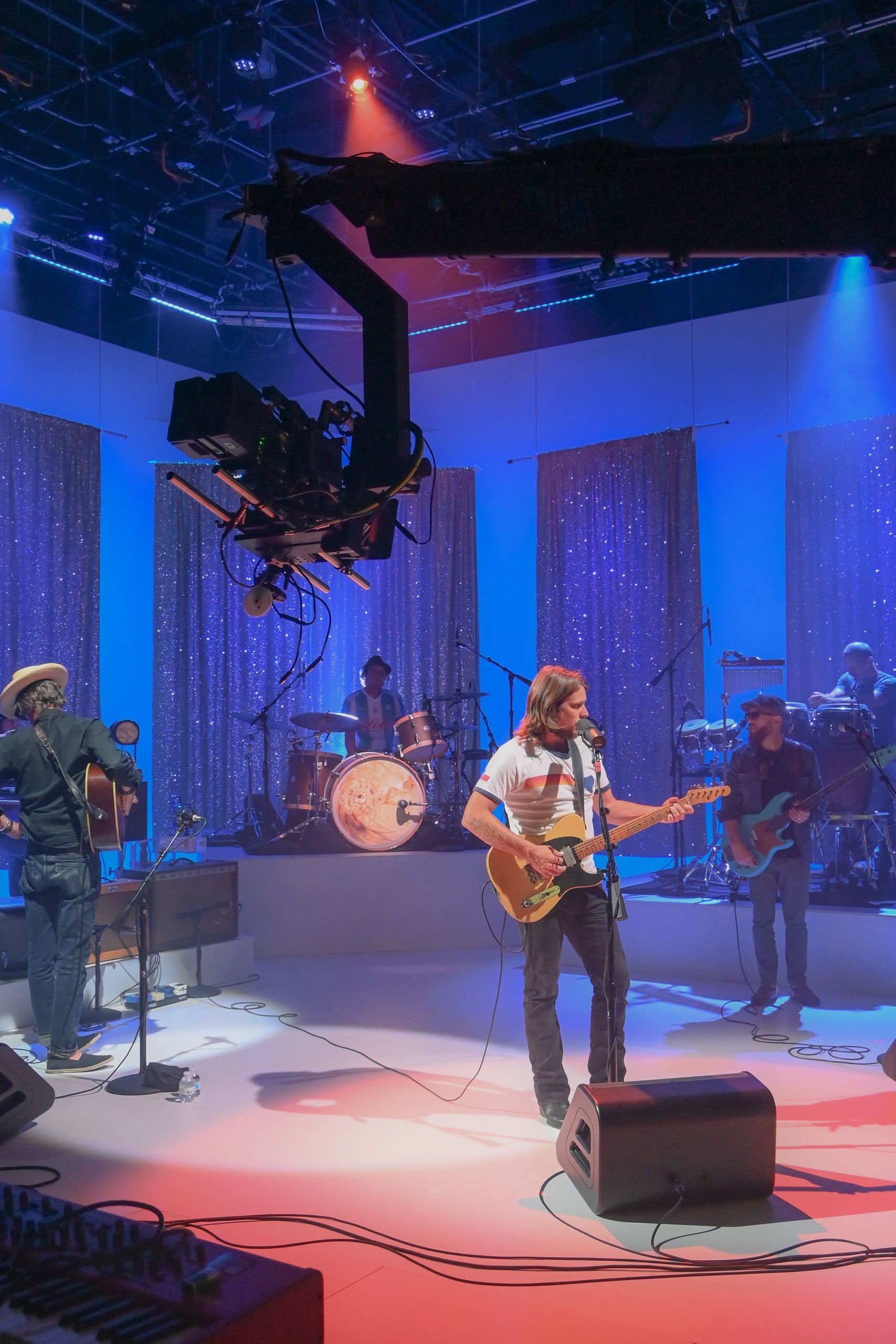 A jib-mounted camera swings over a band playing on a studio stage under blue light