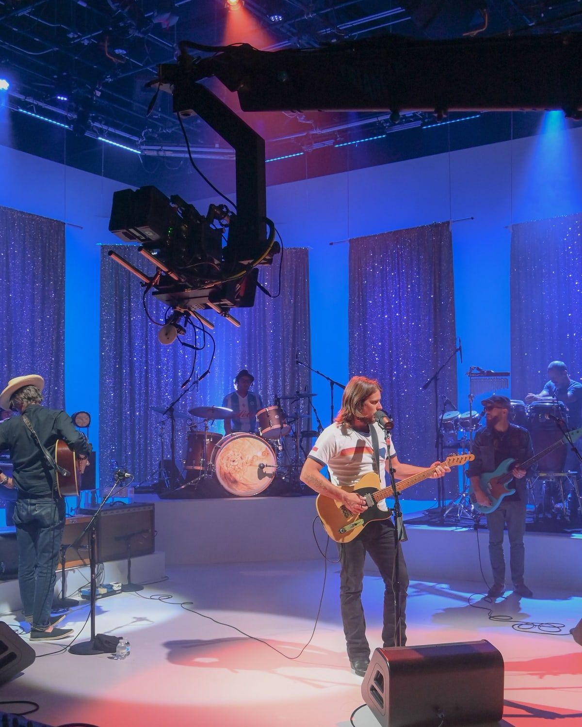 A jib-mounted camera swings over a band playing on a studio stage under blue light