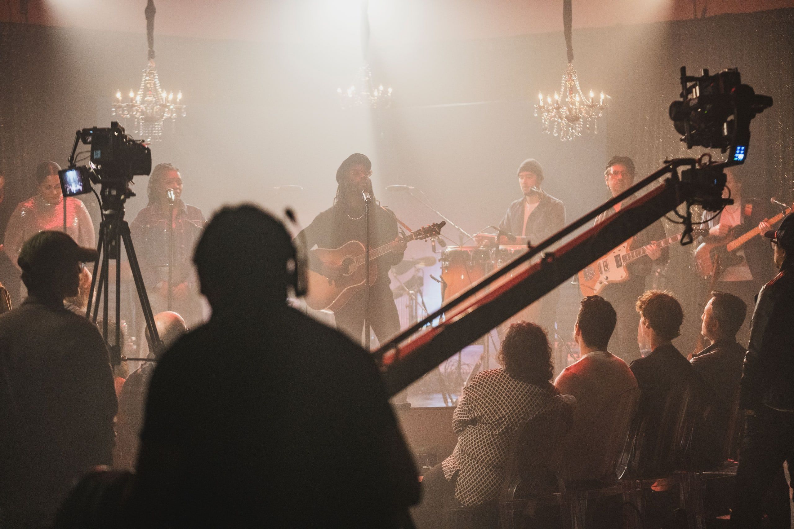 A jib arm swings a camera over a seated crowd as a band plays under chandeliers on a smoky stage