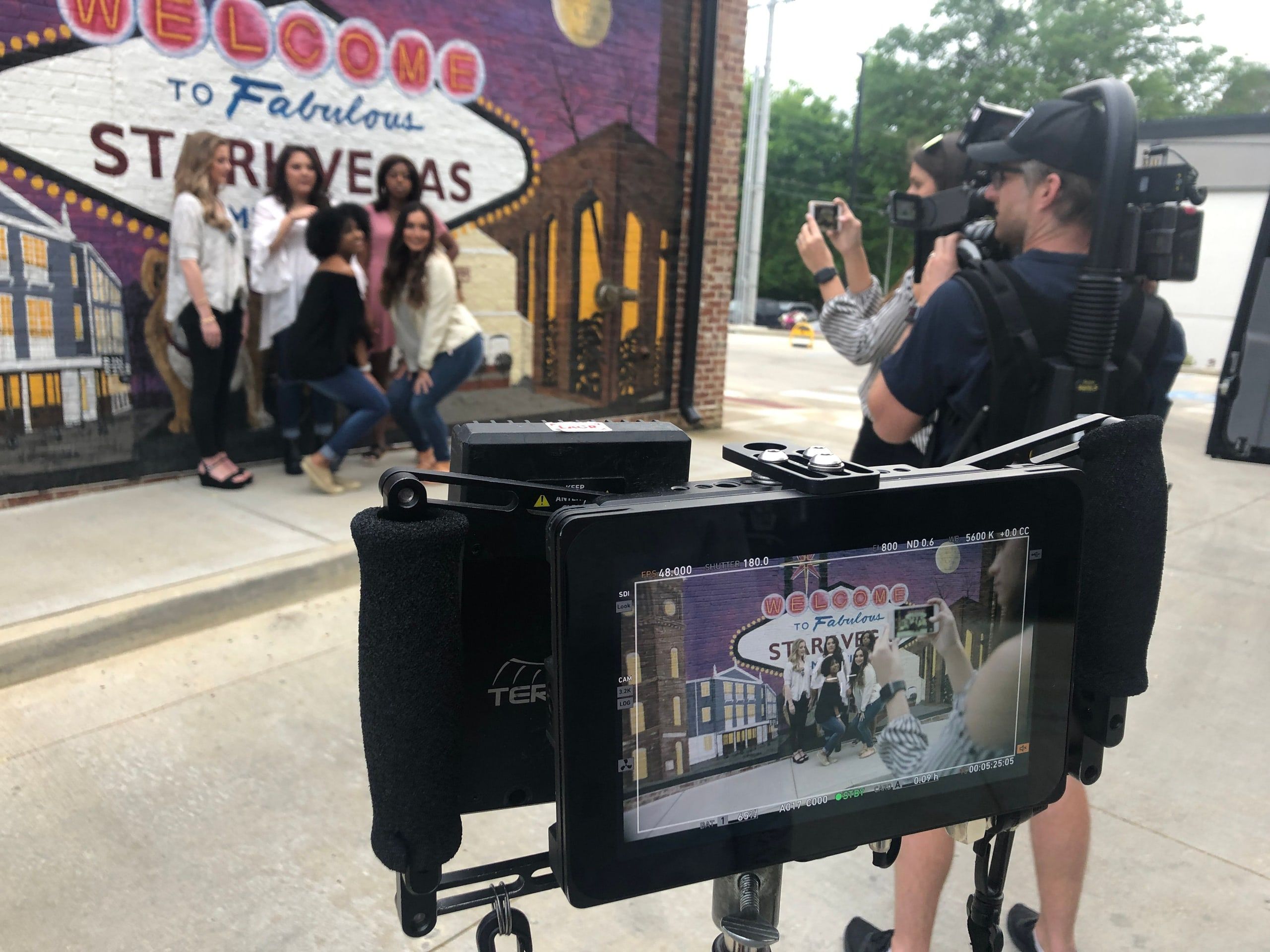 A camera monitor frames five women posing in front of a painted "Welcome to Fabulous" mural while the operator rolls