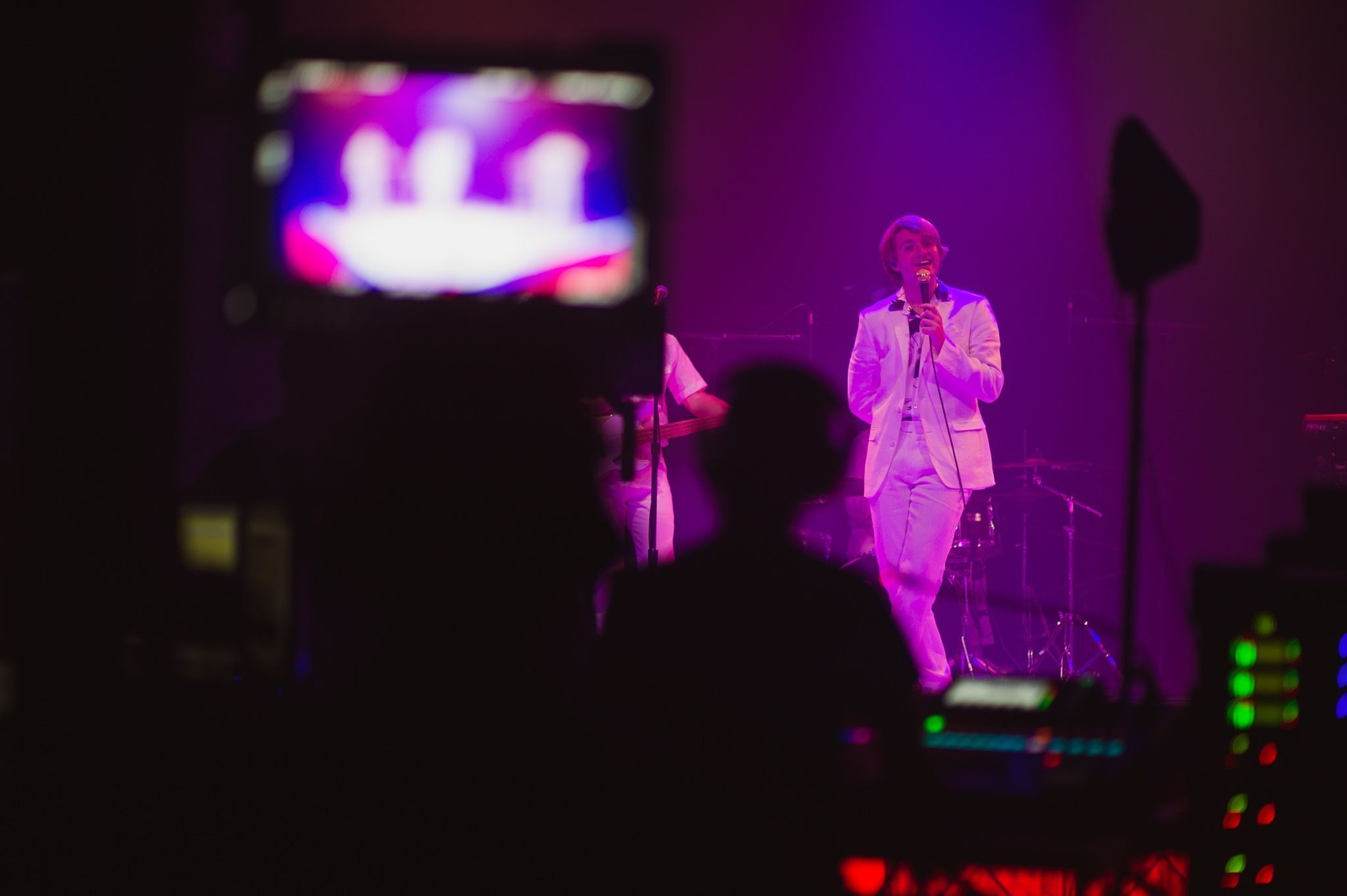A singer in a white suit on a purple-lit stage, seen past a silhouetted camera operator and the video monitor