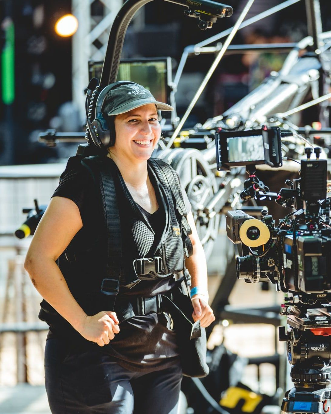A Steadicam operator in her vest and headset, grinning between takes beside the camera at an outdoor stage
